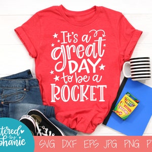 May include: Red t-shirt with white text that reads "It's a great day to be a ROCKET". The shirt is laying on a white surface with a pair of blue jeans, a black and white striped mug, a blue notebook, and a box of crayons.