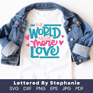 以下が含まれることがあります： 青い文字とピンクの文字で「The World Needs More Love」と書かれた白いTシャツ。テキストはピンクのハートで囲まれています。シャツはデニムジャケットの上に置かれています。