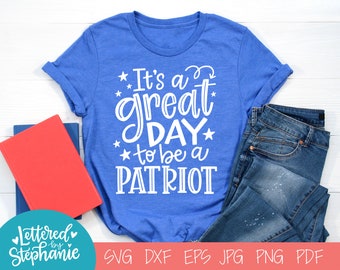 May include: A blue t-shirt with white text that reads "It's a great day to be a PATRIOT". The shirt is on a white surface with a book and jeans.