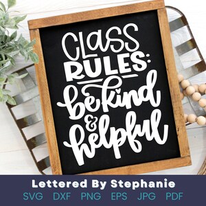 May include: Black chalkboard sign with white text that reads "Class Rules: Be Kind & Helpful". The sign is framed in wood and is sitting on a white wooden surface.