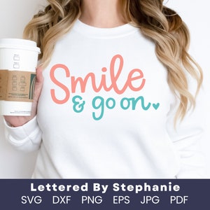 May include: A white sweatshirt with the text "Smile & go on." in pink and blue lettering. The text is in a handwritten style font. The sweatshirt is being held by a person holding a coffee cup.