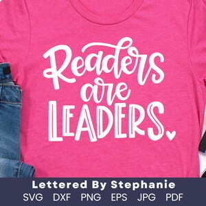 May include: Pink t-shirt with white text that reads "Readers are Leaders."  The text is in a handwritten font.  The shirt is laying on a wooden surface with a pair of blue jeans and a box of crayons.