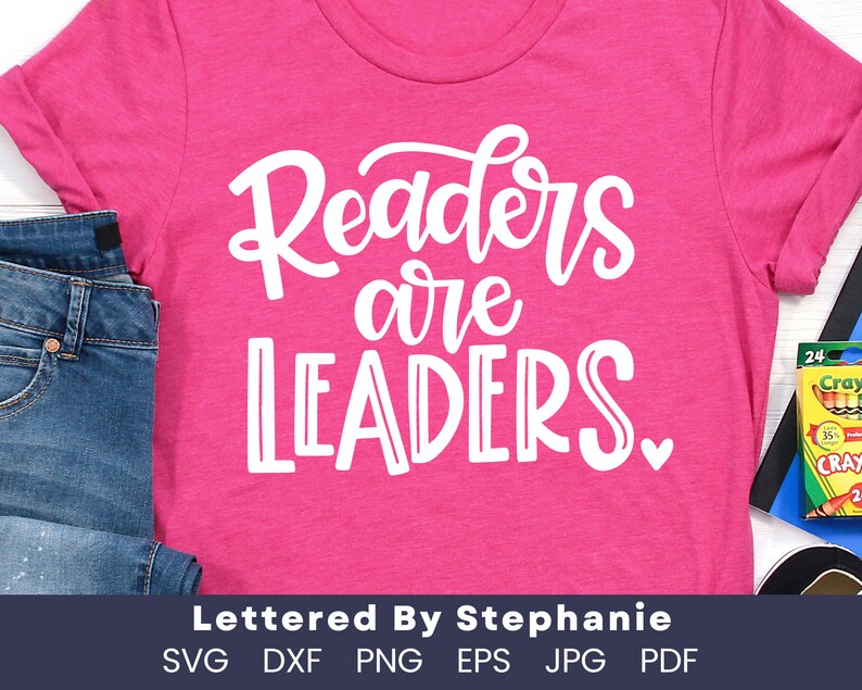 May include: Pink t-shirt with white text that reads "Readers are Leaders."  The text is in a handwritten font.  The shirt is laying on a wooden surface with a pair of blue jeans and a box of crayons.
