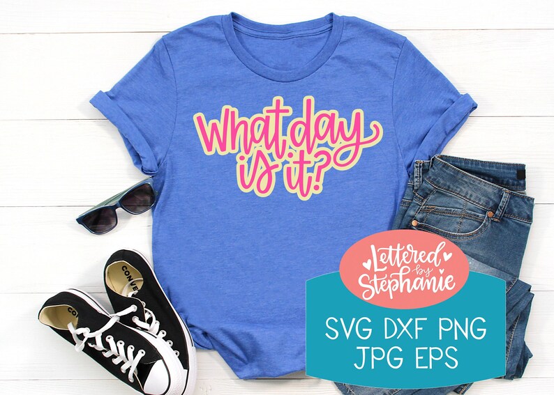 May include: A blue t-shirt with the text "What day is it?" in pink and yellow lettering. The shirt is laying on a white surface with a pair of black sneakers and a pair of sunglasses. A pink and blue oval sticker with the text "Lettered by Stephanie" and the text "SVG DXF PNG JPG EPS" is in the lower right corner of the image.