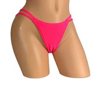Peut inclure: Bas de bikini rose vif avec un dos en string et des détails à double lanière sur les côtés. Le maillot de bain est présenté sur un mannequin sur fond blanc. Le tissu semble lisse et moulant.