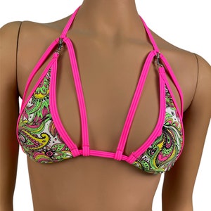 May include: A vibrant bikini top with a paisley pattern in green, yellow, and pink on a white background. The top features bright pink straps and trim, with silver-tone metal connectors. The halter style and strappy design create a bold look.