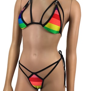 May include: Rainbow-striped bikini set with black trim. The top features a halter neck and cut-out design, with metal clasps. The matching bottom has a strappy design with tie sides. The fabric displays a vibrant spectrum of colors.
