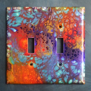 Puede incluir: Una tapa de interruptor de luz doble con un diseño abstracto colorido que presenta remolinos de pintura naranja, roja, azul, púrpura y dorada.