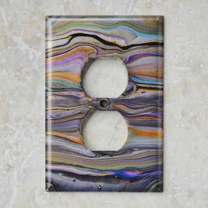 Puede incluir: Cubierta decorativa rectangular para toma de corriente con un diseño abstracto colorido. La cubierta presenta un patrón arremolinado de tonos morados, dorados, azules y grises. Tiene dos aberturas circulares para las tomas y una pequeña abertura central.