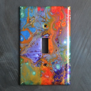 Puede incluir: Cubierta de interruptor de luz decorativa con un diseño abstracto. La cubierta presenta una vibrante mezcla de colores, incluyendo azul, naranja, morado y dorado, creando un efecto marmolado. Tiene una abertura rectangular y dos orificios para tornillos.