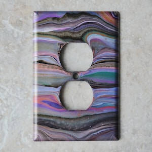 Puede incluir: Cubierta decorativa para enchufe eléctrico con un diseño abstracto y colorido. La cubierta presenta un patrón arremolinado de morados, azules, verdes y marrones. Los dos orificios del enchufe son blancos. La cubierta es rectangular y tiene un acabado brillante.