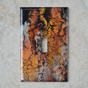May include: A decorative light switch cover with an abstract design. The cover features a rectangular shape with a single switch opening. The artwork showcases a blend of colors, including black, orange, yellow, and silver, creating a unique, artistic appearance.