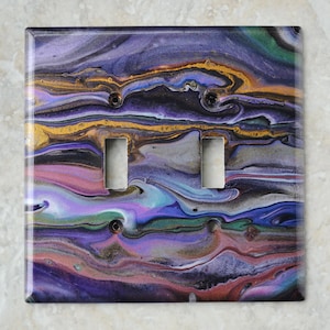 Puede incluir: Cubierta de interruptor de luz doble con un diseño abstracto. La cubierta presenta un patrón arremolinado de tonos morados, dorados, azules y verdes. Tiene dos recortes rectangulares para los interruptores y dos orificios para tornillos.