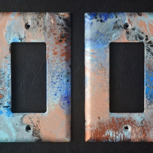 Puede incluir: Dos tapas de interruptor de luz con un diseño abstracto de pintura azul, plata y cobre.