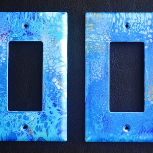 Puede incluir: Dos tapas de interruptor de luz pintadas en azul claro, blanco y dorado con un diseño abstracto en espiral.