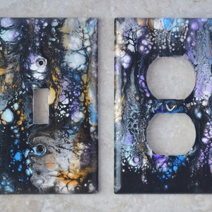 Puede incluir: Dos cubiertas decorativas para interruptores de luz con un diseño abstracto. Las cubiertas presentan un patrón arremolinado de negro, dorado, azul y morado. Una cubierta tiene una abertura para un interruptor, la otra para dos enchufes.