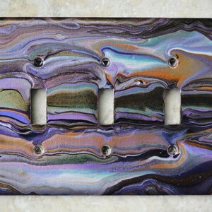 Puede incluir: Placa decorativa para interruptor de luz con un diseño abstracto único. La placa presenta un patrón arremolinado de colores morado, naranja, negro y dorado. Tiene cuatro aberturas para interruptores y está asegurada con tornillos plateados. Un artículo de decoración para el hogar.