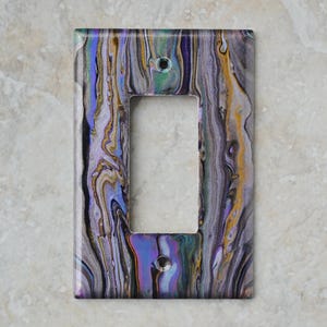 Puede incluir: Una cubierta decorativa para interruptor de luz con un diseño abstracto. La cubierta tiene forma rectangular con una abertura para un solo interruptor. El diseño incorpora patrones fluidos de gris, morado, dorado y azul, creando un efecto marmolado.