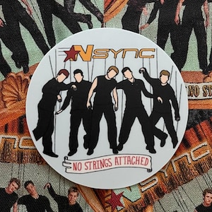 May include: A circular white sticker featuring the boy band *NSYNC. The sticker has a black and white illustration of the band members. The text "NO STRINGS ATTACHED" is on the bottom. The background is a fabric with the band members in black outfits.