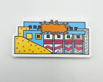 May include: A rectangular sticker featuring a blue building facade with a large orange splatter graphic on top. The design includes a yellow triangular section with pink shapes, vertical red striped panels, and black and white patterned accents.