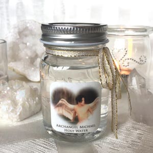 May include: A small glass jar with a silver lid and a gold ribbon tied around it. The jar contains clear liquid and a label that reads "Archangel Michael Holy Water".