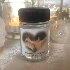 May include: A small clear glass bottle with a black lid and a white label. The label has a picture of an angel with wings and the text "Archangel Michael Holy Water".