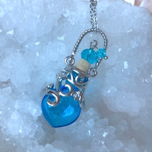 May include: A silver heart-shaped bottle pendant necklace with a blue glass bottle and a cork stopper. The bottle is suspended from a silver chain with a small blue crystal charm.