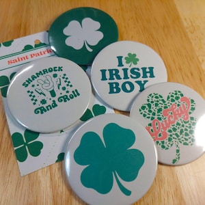 Puede incluir: Cinco botones redondos blancos con diseños de trébol verde. Los botones presentan diferentes diseños, incluyendo un trébol con el texto "Shamrock and Roll", un trébol con el texto "I Irish Boy", un trébol con el texto "Lucky", y dos tréboles.