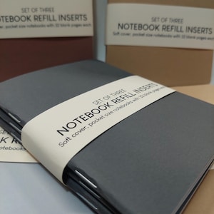 May include: A set of three gray notebook refill inserts with a white label that reads "SET OF THREE NOTEBOOK REFILL INSERTS Soft cover, pocket size notebooks with 32 blank pages each."