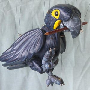 May include: A dark blue wooden puppet of a parrot with a yellow beak and eye. The parrot is holding a wooden pencil in its beak and is suspended by strings.