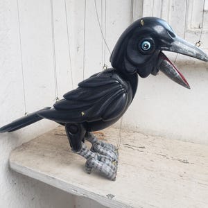 Op de afbeelding: Een zwarte houten kraaienpop met een gedetailleerd ontwerp. De kraai heeft een zilveren snavel, blauwe ogen en een rode binnenkant van de mond. Hij hangt aan touwtjes, wat suggereert dat het een marionet is. De kraai zit op een witte plank.