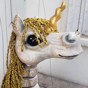 Może przedstawiać: Drewniana marionetka jednorożca z wykończeniem o jasnym usłojeniu drewna, metalicznym złotym rogiem i grzywą z teksturowanej złotej przędzy. Marionetka ma duże ciemne oczy, segmentowaną szyję i cienkie czarne sznurki sterujące przymocowane do metalowych kółek.