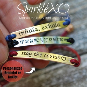 May include: Three gold bracelets with different engraved messages: "inhala, exhala", "42° 38' 24.162" N 70° 52' 56.4744" W", and "stay the course ♥". The bracelets are on a red, blue, and black cord.
