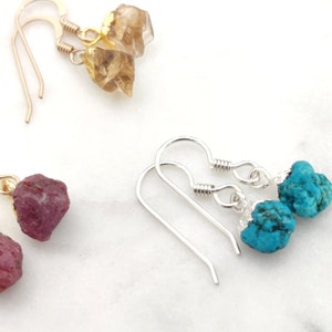 May include: A collection of gemstone earrings. The earrings feature raw, uncut stones in shades of brown, magenta, and turquoise. The earrings are suspended from gold and silver ear wires.