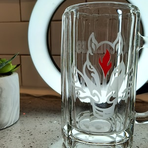May include: A clear glass beer mug with a handle, featuring an etched design of a wolf-like creature with a red flame. The mug is approximately 15 cm tall and sits on a countertop next to a potted succulent.