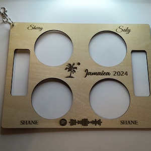May include: A light-coloured wooden drinks holder with four circular cutouts and two rectangular cutouts. The text "Jamaica 2024" and names are engraved on the wood. A Spotify code and a keychain are attached.