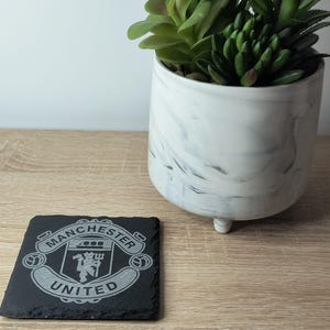 以下が含まれることがあります： 黒い持ち手と「But First, Coffee」のテキストが黒い太字で書かれた白いセラミックマグ。マグカップはぼやけた背景に置かれており、居心地の良い環境を示唆しています。