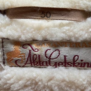 Puede incluir: Una chaqueta de piel de oveja blanca con una etiqueta marr&oacute;n que dice "50" y una etiqueta burdeos con un logotipo de ciervo dorado y el texto "Garanteras Akta Yetskin".