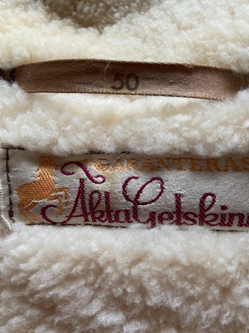 Puede incluir: Una chaqueta de piel de oveja blanca con una etiqueta marr&oacute;n que dice "50" y una etiqueta burdeos con un logotipo de ciervo dorado y el texto "Garanteras Akta Yetskin".