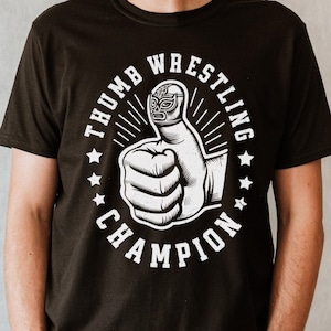 May include: Black t-shirt with a white graphic of a thumb wearing a wrestling mask and the text "Thumb Wrestling Champion".