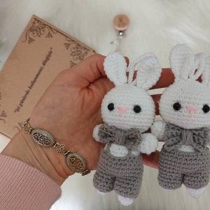 May include: Two crocheted white bunny toys with gray overalls and bow ties. The bunnies are attached to wooden clips with white strings.