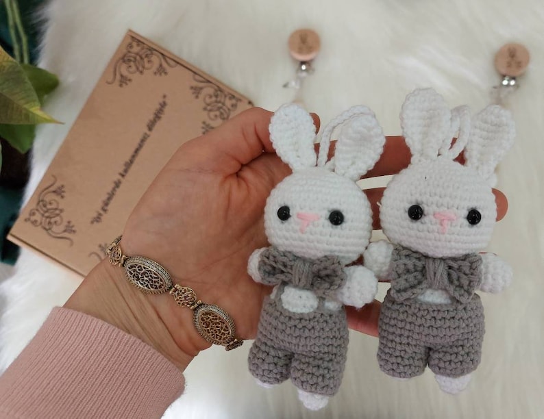 May include: Two crocheted white bunny toys with gray overalls and bow ties. The bunnies are attached to wooden clips with white strings.