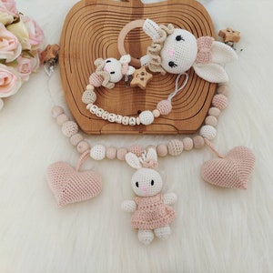 May include: A wooden teething toy with a pink and white crocheted bunny, two pink crocheted hearts, and wooden beads. The toy is personalized with the name "MALIAHA".