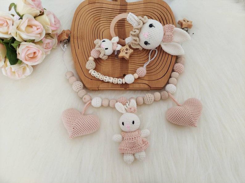 May include: A wooden teething toy with a pink and white crocheted bunny, two pink crocheted hearts, and wooden beads. The toy is personalized with the name "MALIAHA".