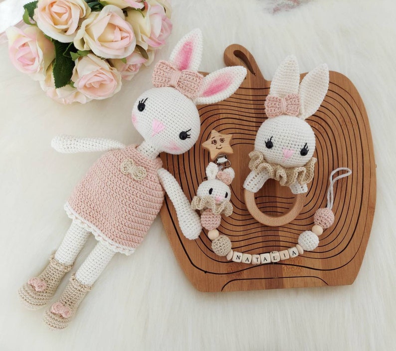 May include: Two crocheted bunny toys with pink accents, one wearing a pink dress and the other with a wooden ring, on a wooden tray with the name "Natalia" spelled out in wooden letters.