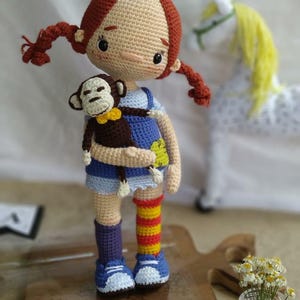 May include: A crocheted doll with red hair, wearing a blue and white dress and holding a brown crocheted monkey. The doll is standing on a wooden cutting board.