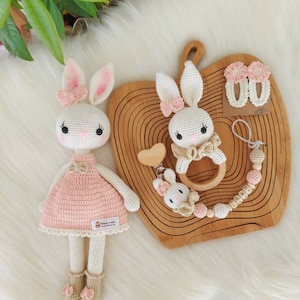 May include: Two crocheted bunny toys, one larger and one smaller, with pink and beige accents. The larger bunny is wearing a pink dress and the smaller bunny is attached to a wooden ring and a wooden bead chain with the name "LEEN" spelled out.