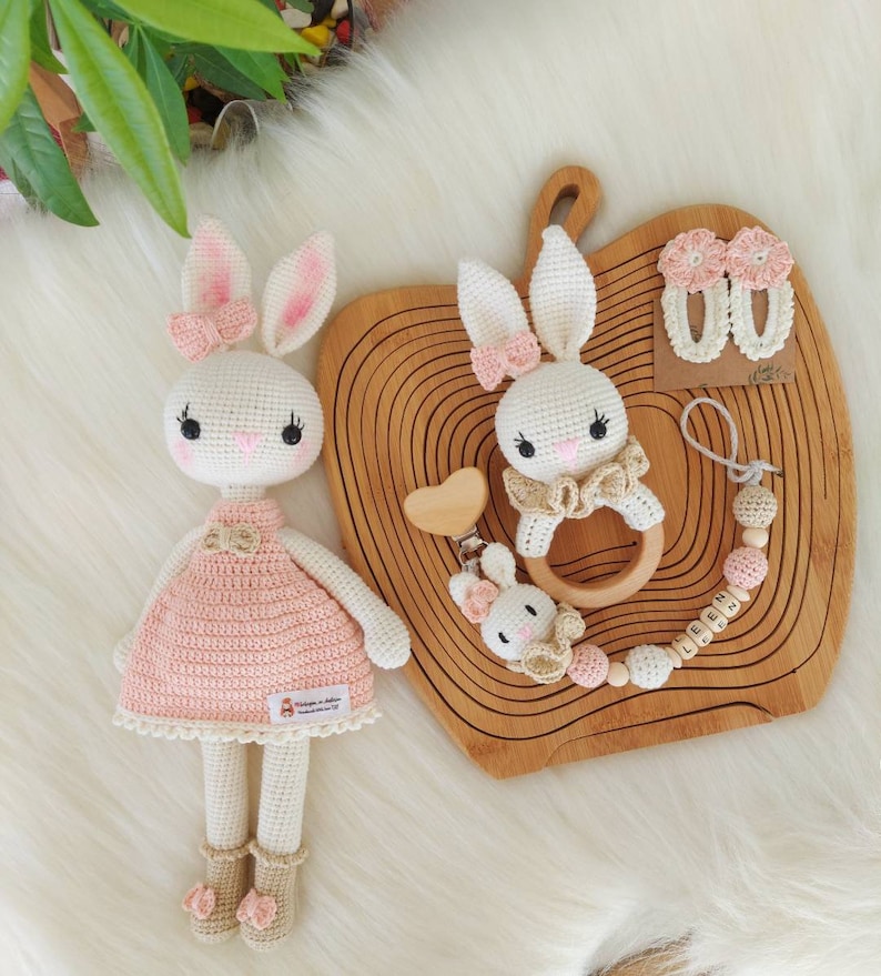 May include: Two crocheted bunny toys, one larger and one smaller, with pink and beige accents. The larger bunny is wearing a pink dress and the smaller bunny is attached to a wooden ring and a wooden bead chain with the name "LEEN" spelled out.