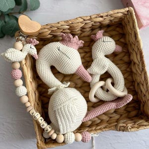 Pode incluir: Um cisne de crochê branco com detalhes rosa e um anel de madeira, um cisne de crochê branco menor com detalhes rosa e um colar de contas de madeira com o nome "ALARA" escrito em blocos de madeira. Os itens estão em uma cesta tecida.