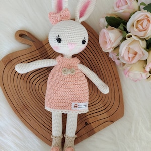 May include: A white crocheted bunny doll wearing a pink dress with a bow. The doll has black eyes and a pink nose. The doll is sitting on a wooden surface.